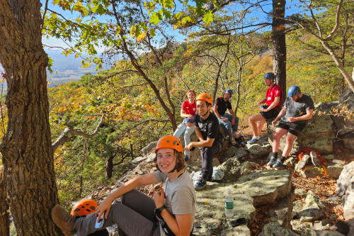 A group of six students wearing hiking gear sit on a rocky overlook surrounded by trees