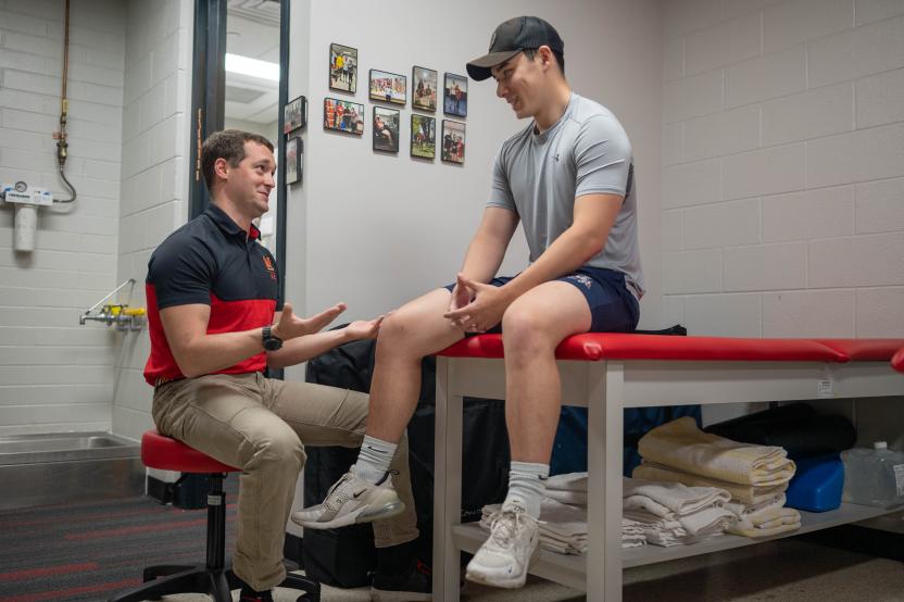 An athletic trainer sits and discusses a student's injury with them as they sit on a treatment table in the clinic.