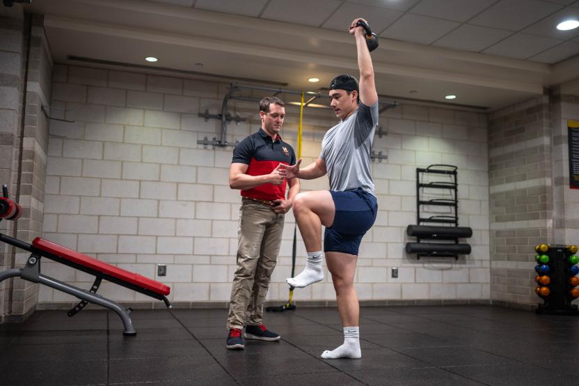 An athletic trainer looks on as a student performs a kettle bell exercise in front of them.