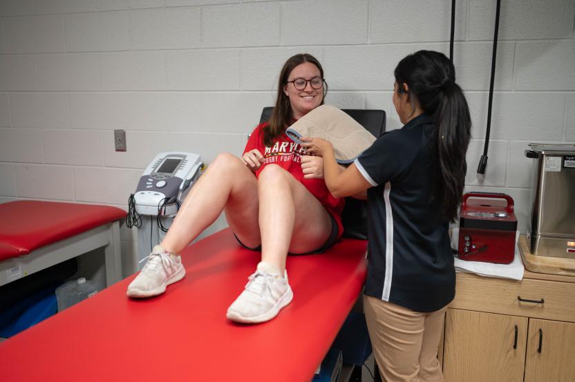 An athletic trainer places a hot pack on a student's shoulder as they sit on a treatment table in the clinic.