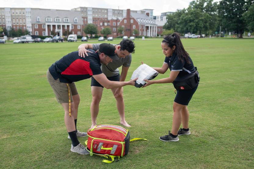 Two athletic trainers stand on a field, assisting a hunched over student.