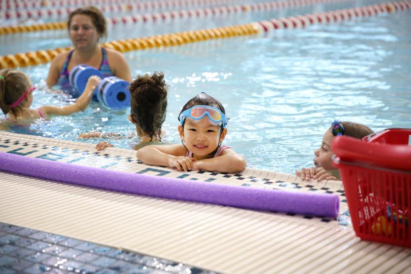 a small child with their arms up on the side of the pool. There is a purple pool noodle in the foreground