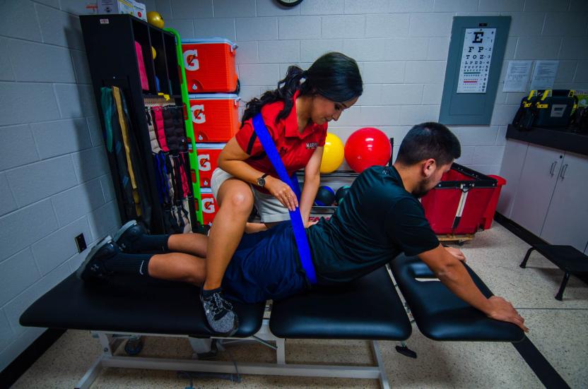 An athletic trainer mobilizes a patient's back with a blue belt as they lay prone on a treatment table in the athletic training clinic.