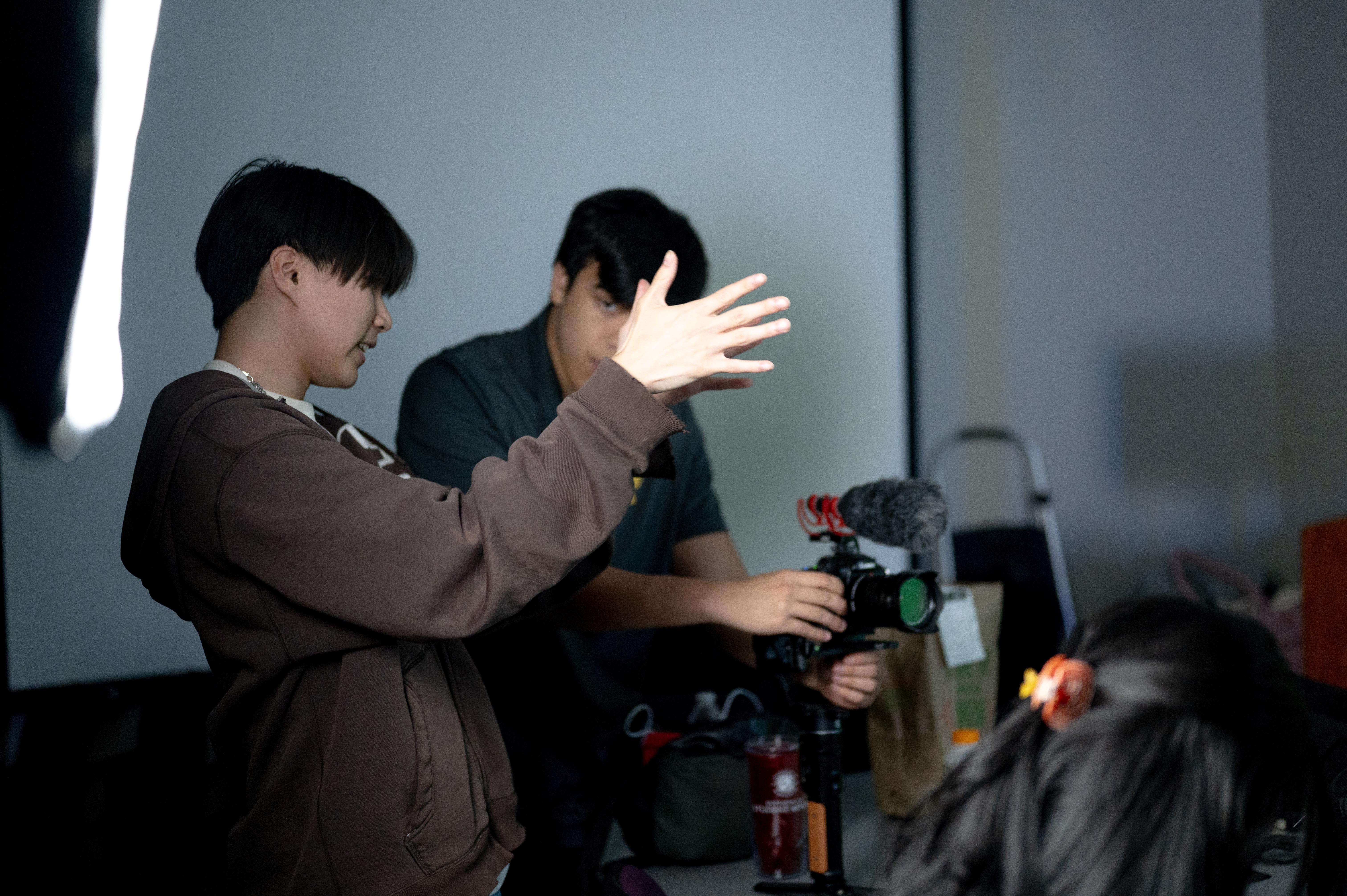 Two content team staff, one holding a camera and the other gesturing with his hands how to frame the camera. 
